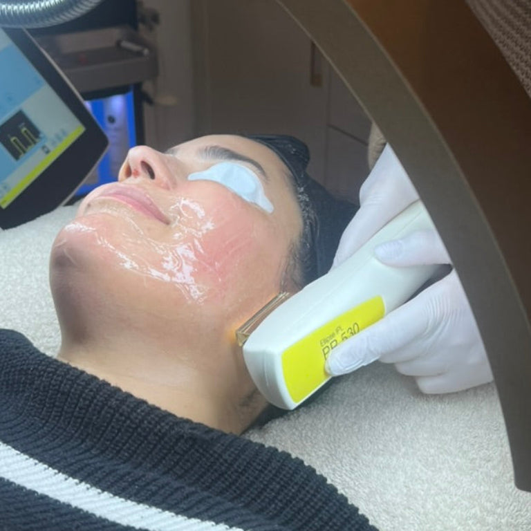 A person lying down during a facial treatment, with a gloved hand holding a handheld device against the cheek. A large round mirror reflects the scene, showing a clinic interior with screens and equipment in the background.