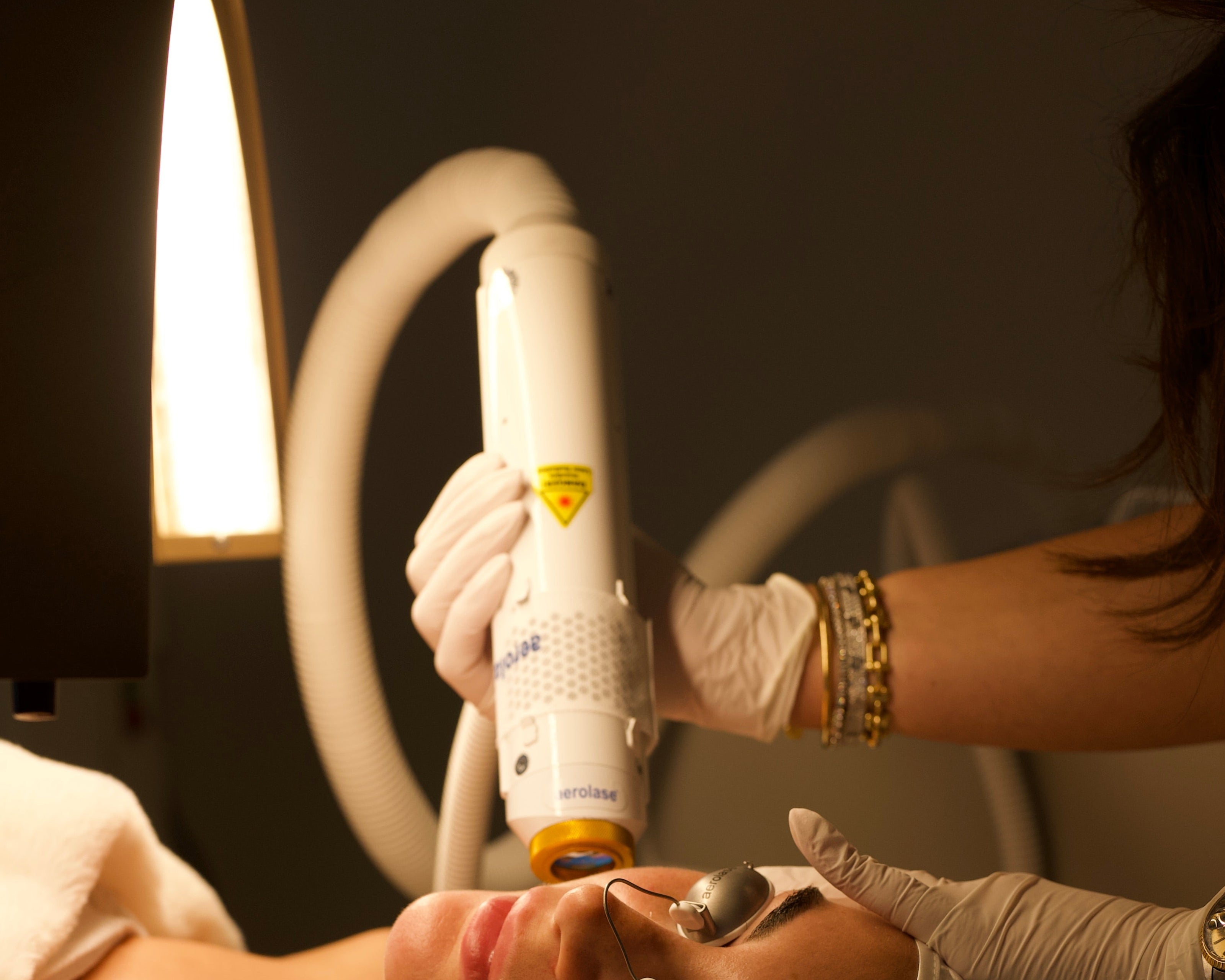 A woman lies on a treatment bed while a practitioner in white gloves uses a handheld white and yellow device on her face, with warm lighting in a spa-like setting.