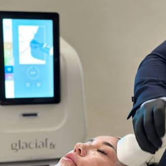 A person lying down with a white headband while another person in a dark blue sleeve and black glove applies a white cosmetic or treatment device to their face. A white and gray glaciaf6 device with a touchscreen display is visible in the background.
