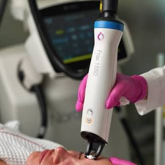 A close-up of a skincare treatment in progress, showing a person lying down while a gloved hand holds a white and blue FlexiLift 1500 device against their face. Another gloved hand gently holds the face open.