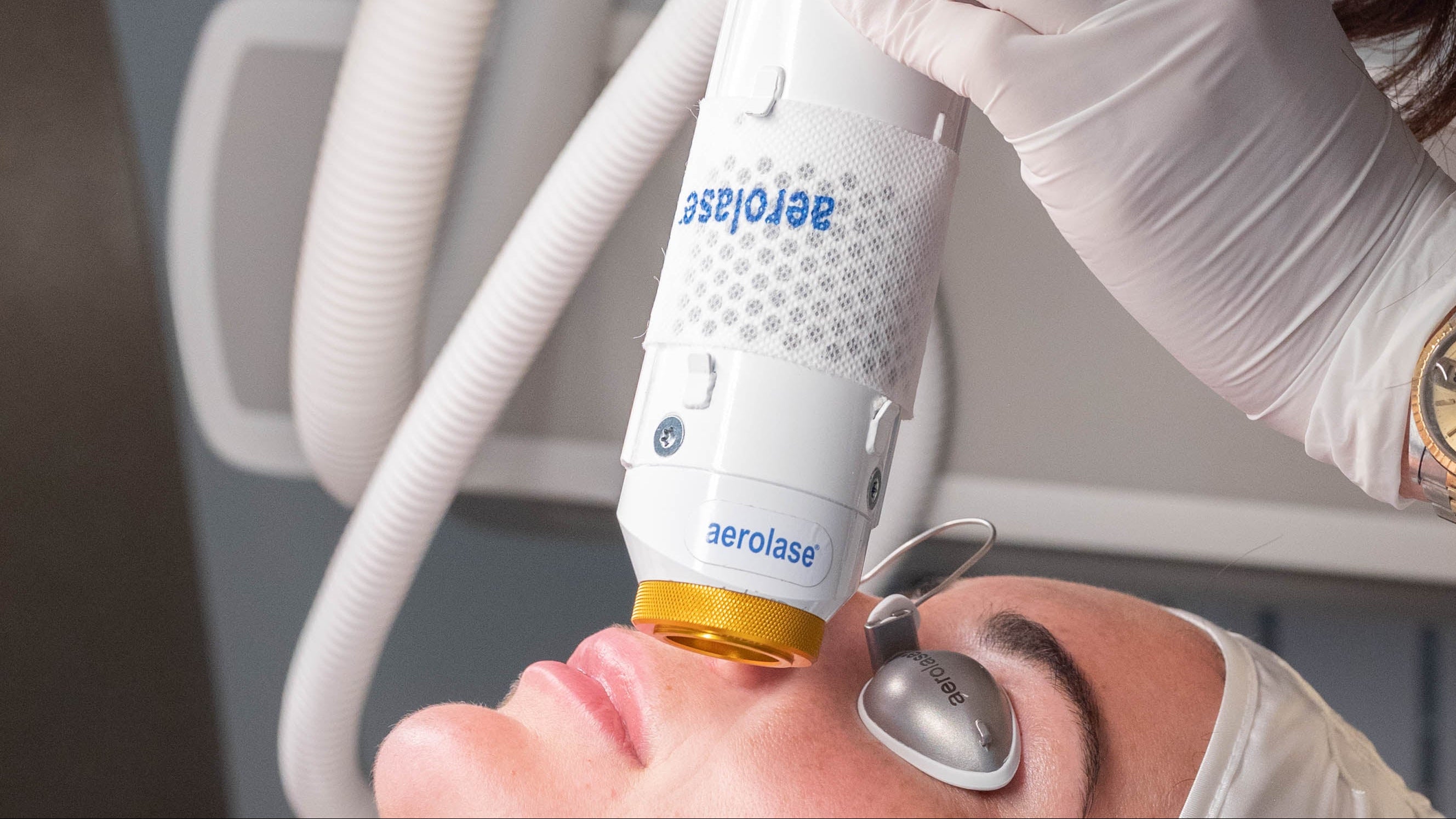 A close-up of a beauty therapist using a white and gold handheld facial treatment device on a client's face, with the therapist wearing white gloves and a cream-colored uniform.